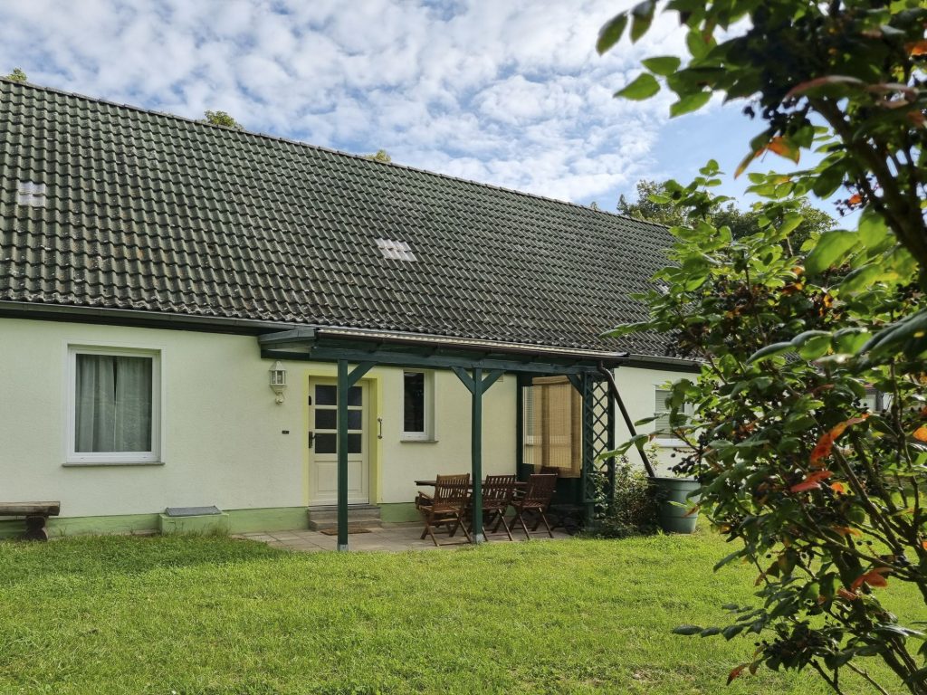 Blick vom Garten auf die Südseite des Hauses mit überdachter Terrasse und Eingang. Im Vordergrund Rondell-Bepflanzung.